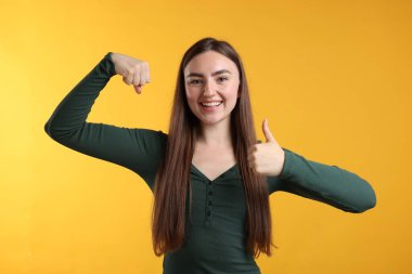 Kuvvet. Sarı arka planda bisepslerini ve başparmaklarını gösteren mutlu kadın.