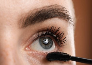 Genç bir kadın kahverengi arka planda kirpiklerine rimel sürüyor, yakın plan.