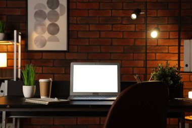 Laptop, paket kâğıt bardak, ev bitkileri ve tuğla duvarın yanındaki siyah masada kırtasiye malzemeleri.