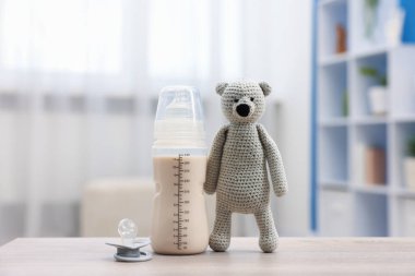 Sütlü bebek şişesi, emzik ve oyuncak ayı masada.