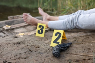 Adli tıp uzmanlığı. Cesedin bulunduğu suç mahalli, gölün yakınında işaretler ve silah, seçici bir odak noktası.