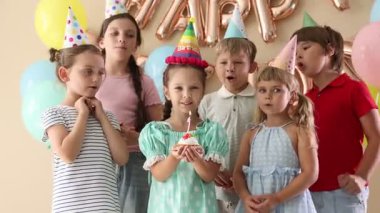 Doğum günü kutlaması. Tatlı küçük kız kekin üzerine mum üflüyor, arkadaşları ve bej arka planda şenlik dekoru.
