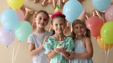 Doğum günü kutlaması. Tatlı küçük kız kekin üzerine mum üflüyor, arkadaşları ve bej arka planda şenlik dekoru.