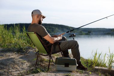 Yazın sandalyede oturan ve göl kenarında balık tutan oltalı balıkçı.