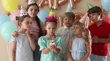 Doğum günü kutlaması. Tatlı küçük kız kekin üzerine mum üflüyor, arkadaşları ve bej arka planda şenlik dekoru.
