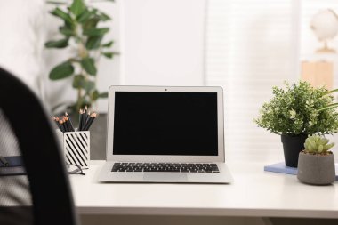 Ofisteki beyaz masada dizüstü bilgisayarı, kırtasiyesi ve çiçek dekoru olan şık bir iş yeri.