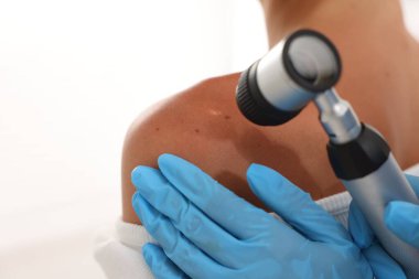 Doktor beyaz arka planda dermatoskopla genç kadının benini inceliyor.