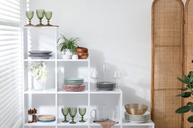 Beyaz duvarın yanında farklı bulaşık takımları, gözlük ve çiçek dekoru olan raf ünitesi.