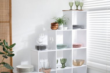 Beyaz duvarın yanında farklı bulaşık takımları, gözlük ve çiçek dekoru olan raf ünitesi.