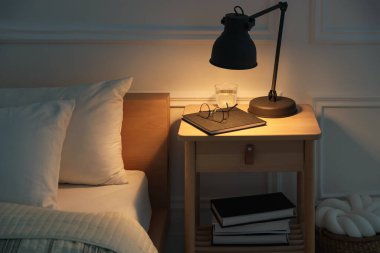 İçinde lamba, defter, bardaklar ve yatağın yanında bir bardak su olan komodin.