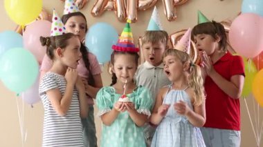 Doğum günü kutlaması. Tatlı küçük kız kekin üzerine mum üflüyor, arkadaşları ve bej arka planda şenlik dekoru.