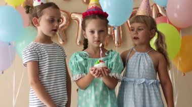 Doğum günü kutlaması. Tatlı küçük kız kekin üzerine mum üflüyor, arkadaşları ve bej arka planda şenlik dekoru.