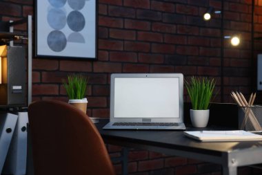 Laptop, ev bitkileri ve kırtasiye malzemeleri tuğla duvarın yanındaki siyah masada.