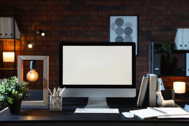 Bilgisayar, ev bitkisi, lamba ve kırtasiye malzemesi tuğla duvarın yanındaki siyah masada.