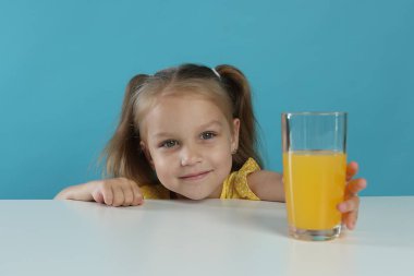 Beyaz masada açık mavi arka planda bir bardak portakal suyuna uzanan tatlı küçük kız.