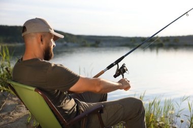 Yazın sandalyede oturan ve göl kenarında balık tutan oltalı balıkçı.