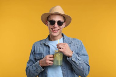 Sarı arka planda limonata kavanozu olan şapkalı ve güneş gözlüklü mutlu bir adam. Tazeleyici içecek.