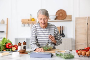 Mutfaktaki beyaz mermer masada yemek pişirirken tablet kullanan yaşlı kadın.