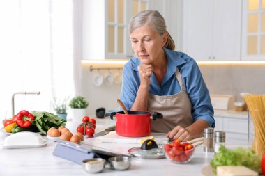 Mutfaktaki beyaz mermer masada yemek pişirirken tablet kullanan yaşlı kadın.