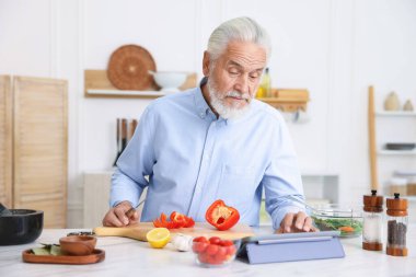Mutfaktaki beyaz mermer masada yemek pişirirken tablet kullanan yaşlı adam.