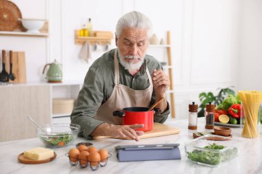 Mutfaktaki beyaz mermer masada yemek pişirirken tablet kullanan yaşlı adam.