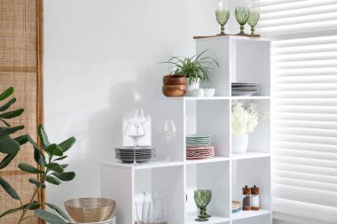 Beyaz duvarın yanında farklı bulaşık takımları, gözlük ve çiçek dekoru olan raf ünitesi.