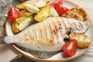 Hafif ahşap masada patates, domates ve limon dilimleriyle servis edilen kızarmış balık, yakın plan. Ev yapımı deniz ürünleri yemeği.