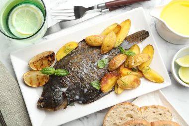 Beyaz mermer masada patates, fesleğen ve limon dilimleriyle servis edilen kızarmış balık. Ev yapımı deniz ürünleri yemeği.