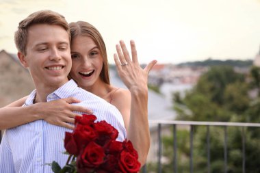 Evlilik teklifi. Nişan yüzüğünü gösteren mutlu bir kadın ve nişanlısı balkonda gül buketi tutuyor. Metin için boşluk