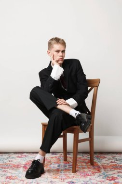 Şık takım elbiseli yakışıklı genç adam beyaz arka planda sandalyede oturuyor.