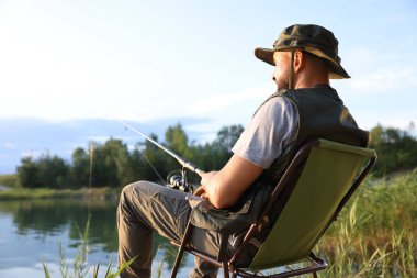 Yazın sandalyede oturan ve göl kenarında balık tutan oltalı balıkçı.