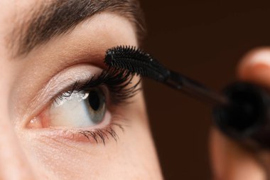 Genç bir kadın kahverengi arka planda kirpiklerine rimel sürüyor, yakın plan.