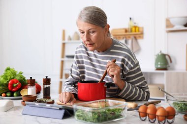 Mutfaktaki beyaz mermer masada yemek pişirirken tablet kullanan yaşlı kadın.