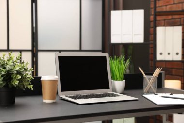 Laptop, paket kâğıt bardak, ev bitkileri ve kırtasiye malzemeleri evdeki siyah masada.