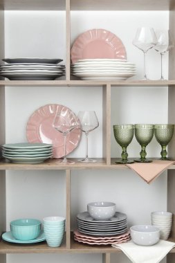 Beyaz duvarın yanında farklı bulaşık takımları ve gözlüklerle raf ünitesi.
