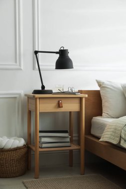 İçinde lamba, defter, gözlük, kitap ve yatağın yanında bir bardak su olan komodin.