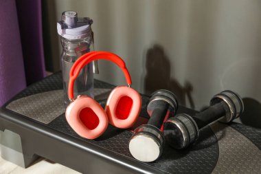 Kablosuz kulaklıklar, bir şişe su ve spor salonunda merdivenlerde duran dambıllar.