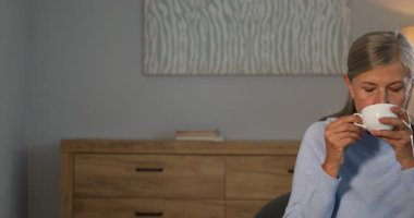Yaşlı kadın evdeki kanepede çay içiyor. Kamera sağa hareket ediyor
