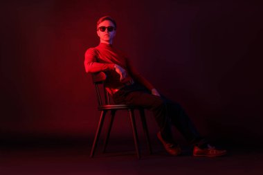 Güneş gözlüğü takan yakışıklı adam, karanlık arka planda, neon ışıklarda, sandalyede oturuyor.