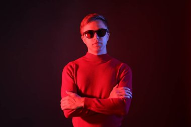 Güneş gözlüğü takan yakışıklı bir adamın portresi. Koyu renk arka planda çapraz kollu, neon ışıklı.