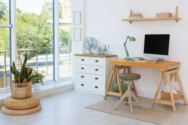 Ev çalışma alanı. Modern bilgisayar ve kırtasiye malzemeleri pencerenin yanındaki ahşap masada.
