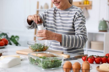 Yemek pişirme süreci. Üst düzey kadın, mutfaktaki beyaz mermer masada salataya baharat ekliyor.