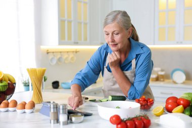Mutfaktaki beyaz mermer masada yemek pişirirken tablet kullanan yaşlı kadın.