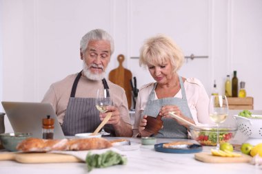 Laptoplu yaşlı çift birlikte yemek pişiriyor ve içeride beyaz şarap içiyor.