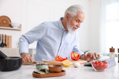 Mutfaktaki beyaz mermer masada yemek pişirirken tablet kullanan yaşlı adam.