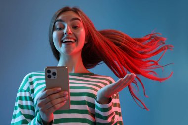 Güzel, uzun saçlı mutlu kadın mavi arka planda, düşük açılı, neon ışıklarda akıllı telefon kullanıyor.