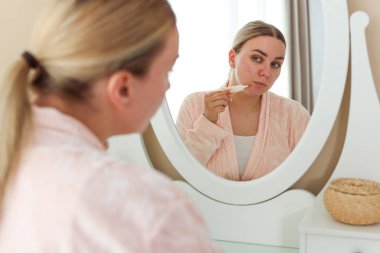 Kadın evin içinde aynanın yanında yüzüne krem sürüyor. Sivilce tedavisi