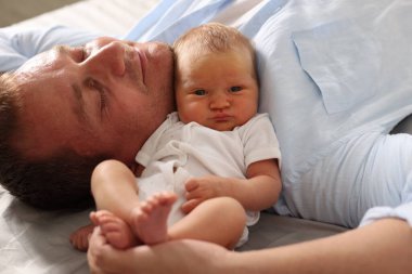 Baba, yeni doğmuş bebeğiyle evde yatakta uzanıyor, yakın plan.