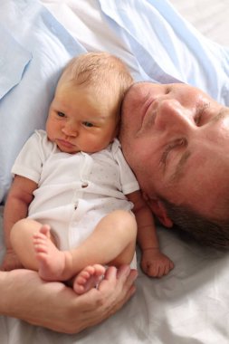 Baba, yeni doğmuş bebeğiyle evde yatakta uzanıyor, yakın plan.
