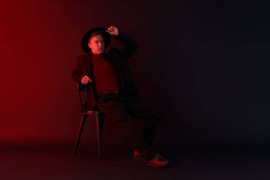 Yakışıklı, takım elbiseli ve şapkalı bir adam, karanlık arka planda, neon ışıklarda oturuyor.